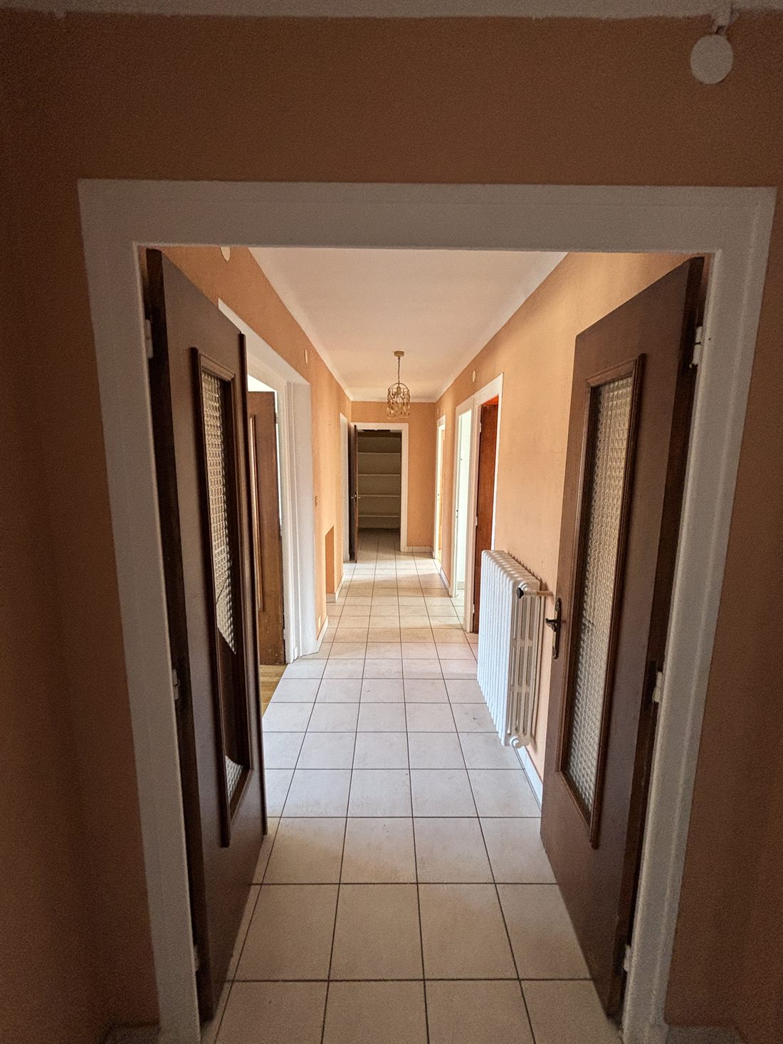 Le couloir avant les travaux, vu depuis une double porte vitrée : murs orangés, carrelage blanc, portes en bois foncé et radiateur en fonte