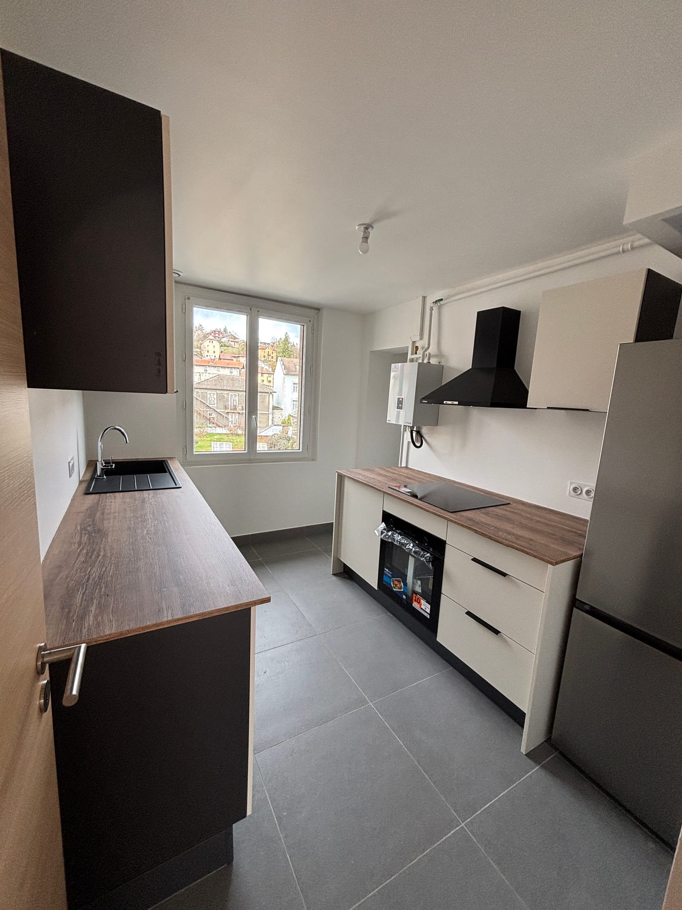 La cuisine après les travaux : meubles neufs, plan de travail bois, plaque et four encastrés, hotte noire, grand carrelage gris et fenêtre sur la ville