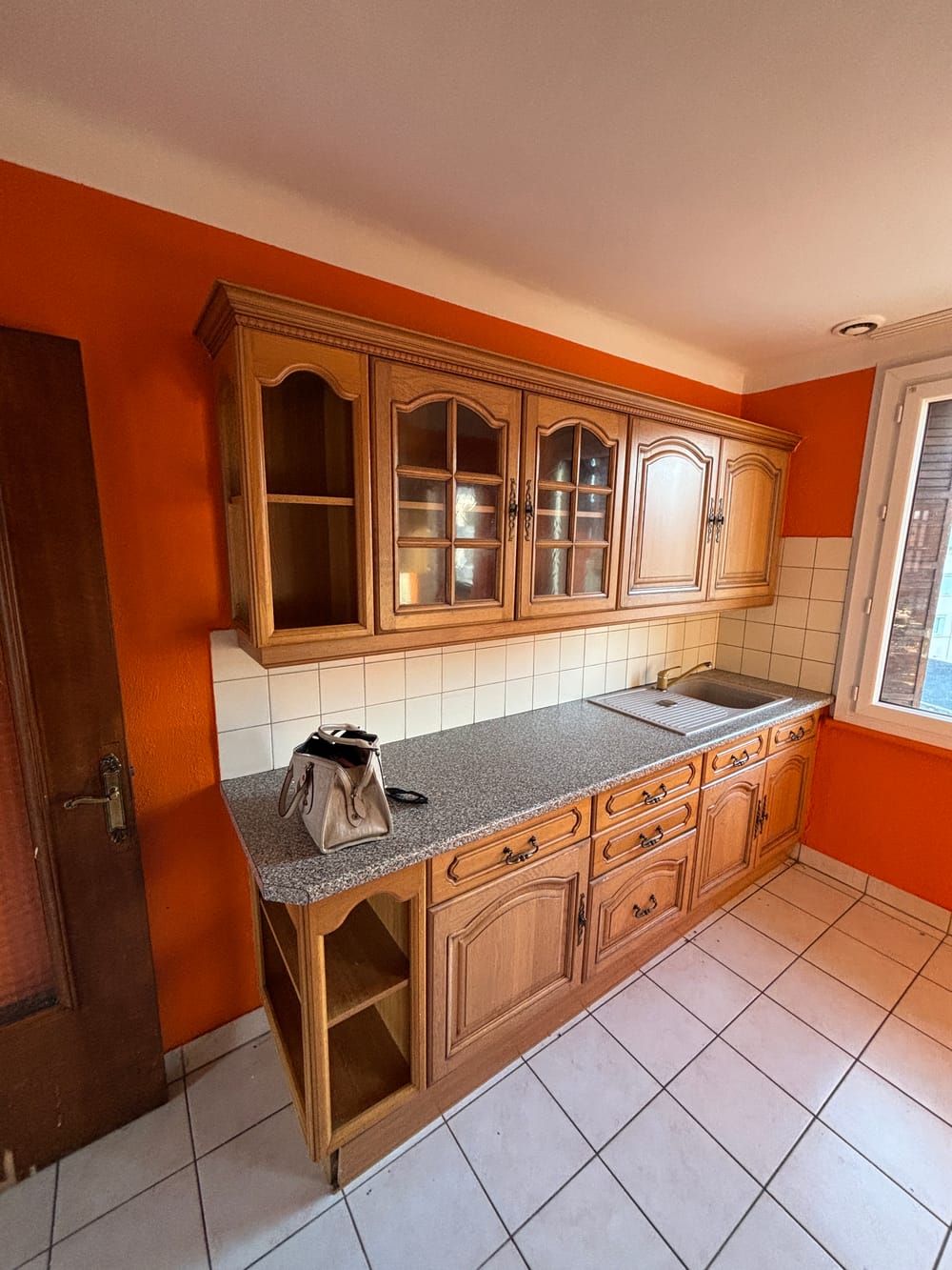 La cuisine avant les travaux : meubles en chêne à portes vitrées, plan de travail granité, murs orange et carrelage blanc