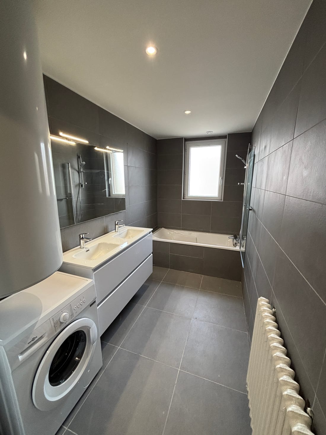 La salle de bains après les travaux : carrelage gris grand format, double vasque suspendue, miroir éclairé et baignoire sous la fenêtre