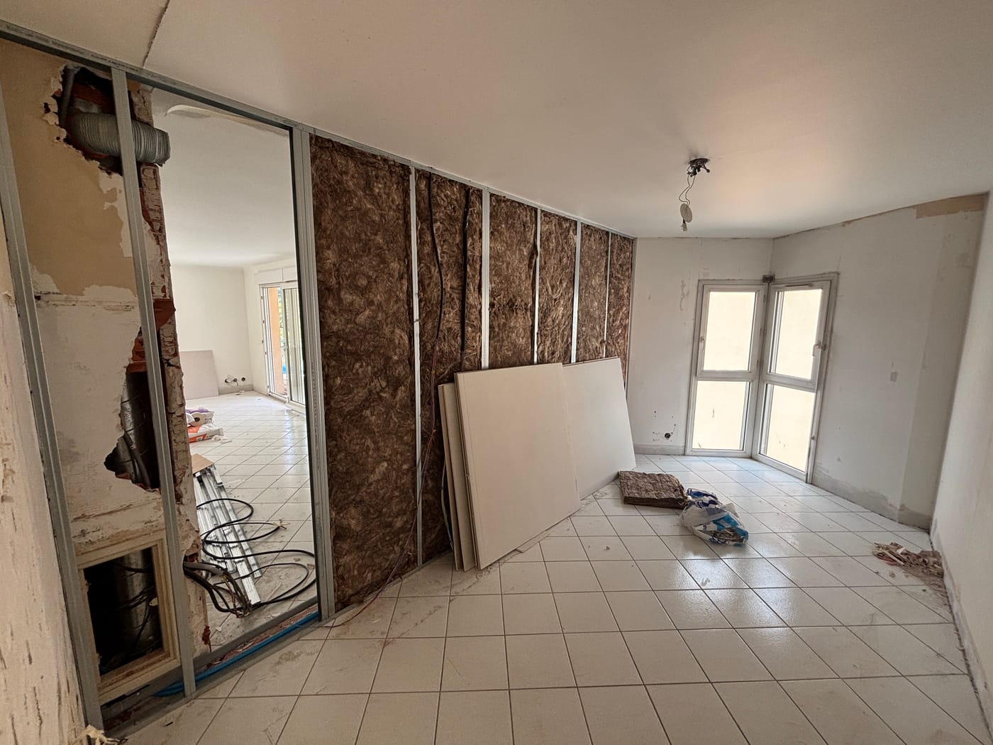 Une pièce pendant les travaux : ossature métallique garnie d'isolant, plaques de plâtre posées contre le mur, ancien carrelage blanc encore en place