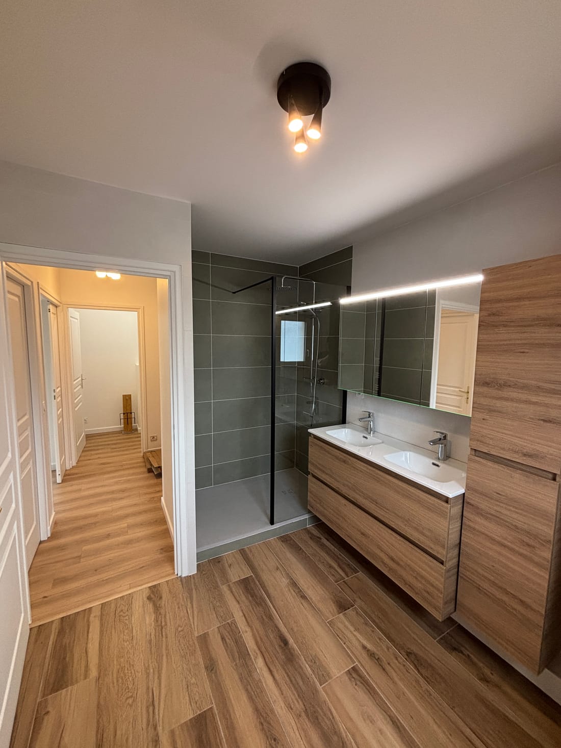 Salle de bains du projet Le Nautile : douche à paroi vitrée, carrelage gris, meuble double vasque et colonne en bois, porte ouverte sur le couloir