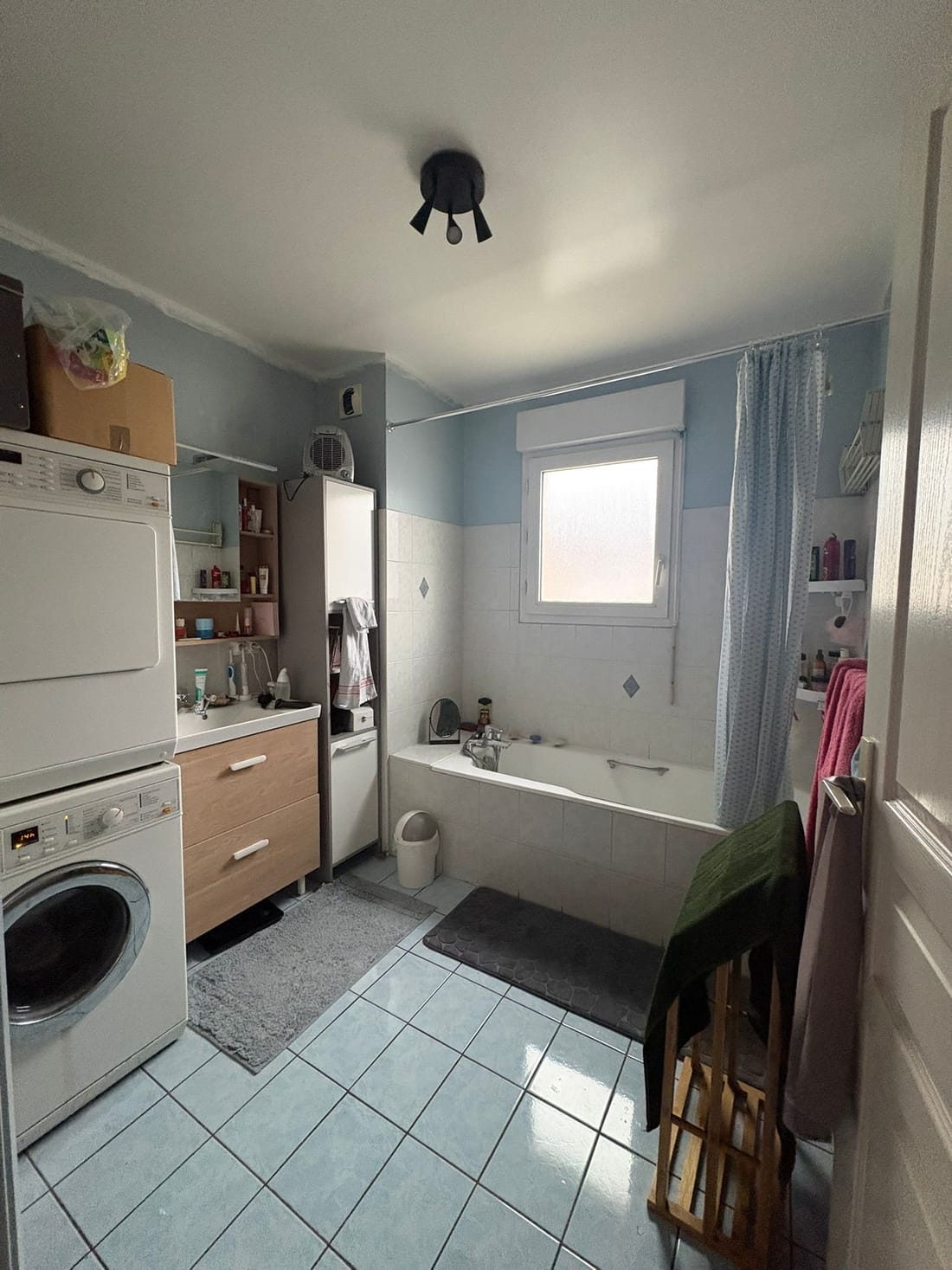 La salle de bains avant les travaux : baignoire sous la fenêtre, faïence blanche, murs bleu clair et carrelage bleu au sol