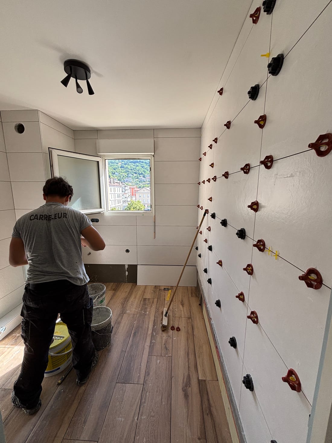 La salle de bains pendant les travaux : un carreleur pose une faïence grand format calée par des croisillons, sol aspect bois déjà posé